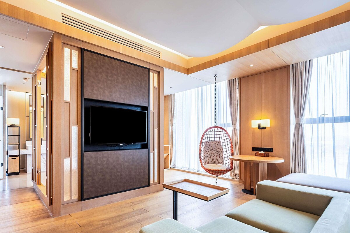 A modern hotel room with a large flat-screen TV mounted on a wooden panel, a hanging chair, a small round table, and a sofa.