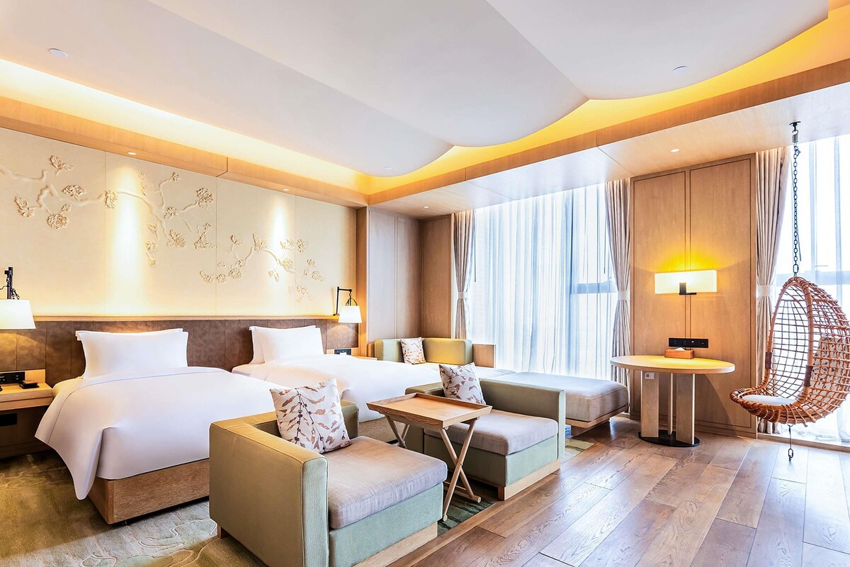 A hotel room with two beds, a sofa, a small table, a hanging chair, and a large window with curtains.