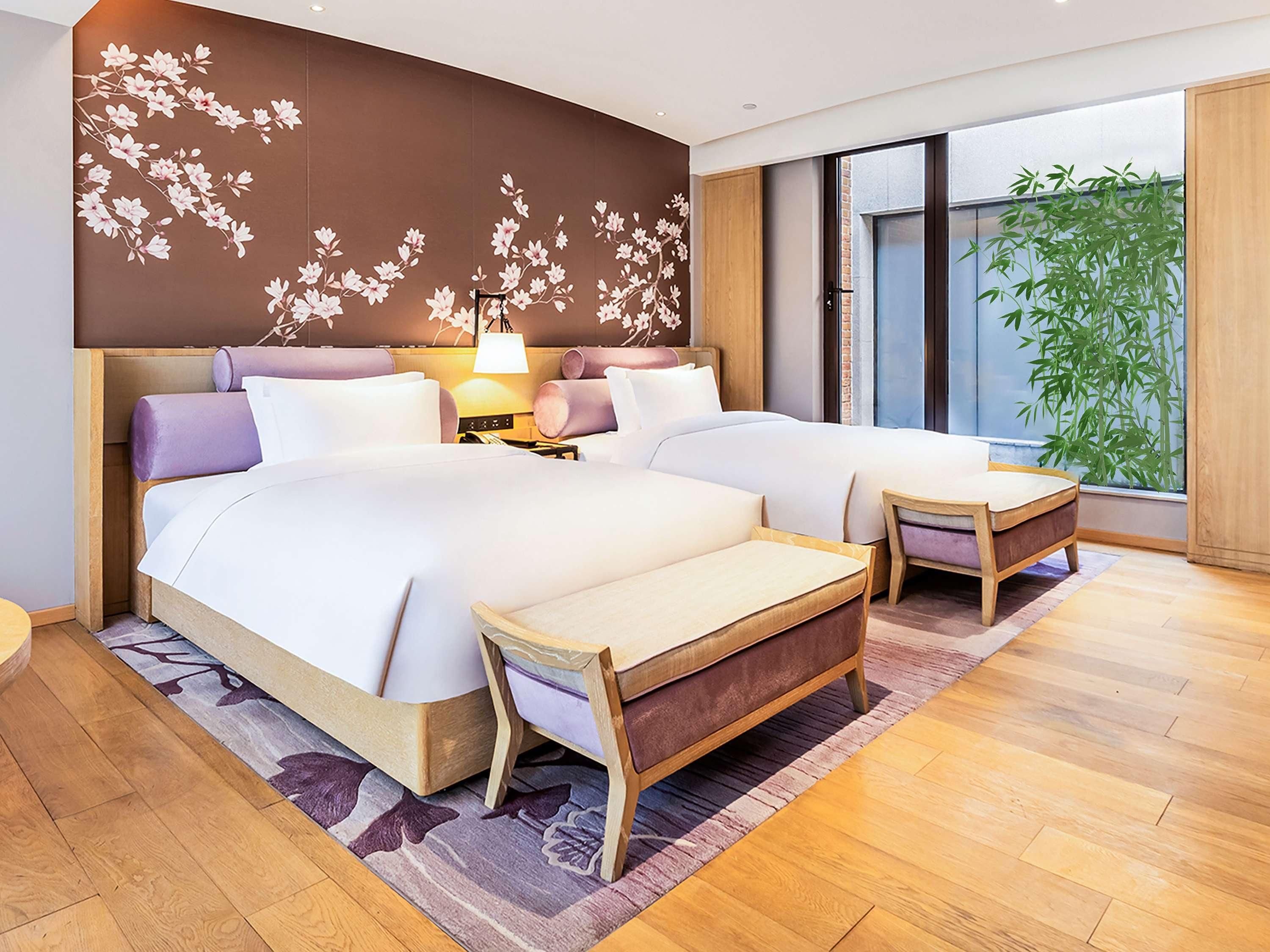 Una habitación de hotel moderna con dos camas, un cabecero de madera, una mesita de noche con lámpara, un ventanal con vistas a bambú y una pared decorativa con motivos florales.