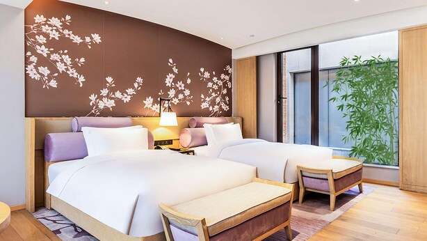 A modern hotel room with two beds, a wooden headboard, a nightstand with a lamp, a large window with a view of bamboo, and a decorative wall with floral patterns. - Swissotel Hot Spring Hotel Xi'an Xinxing (Xi'an)