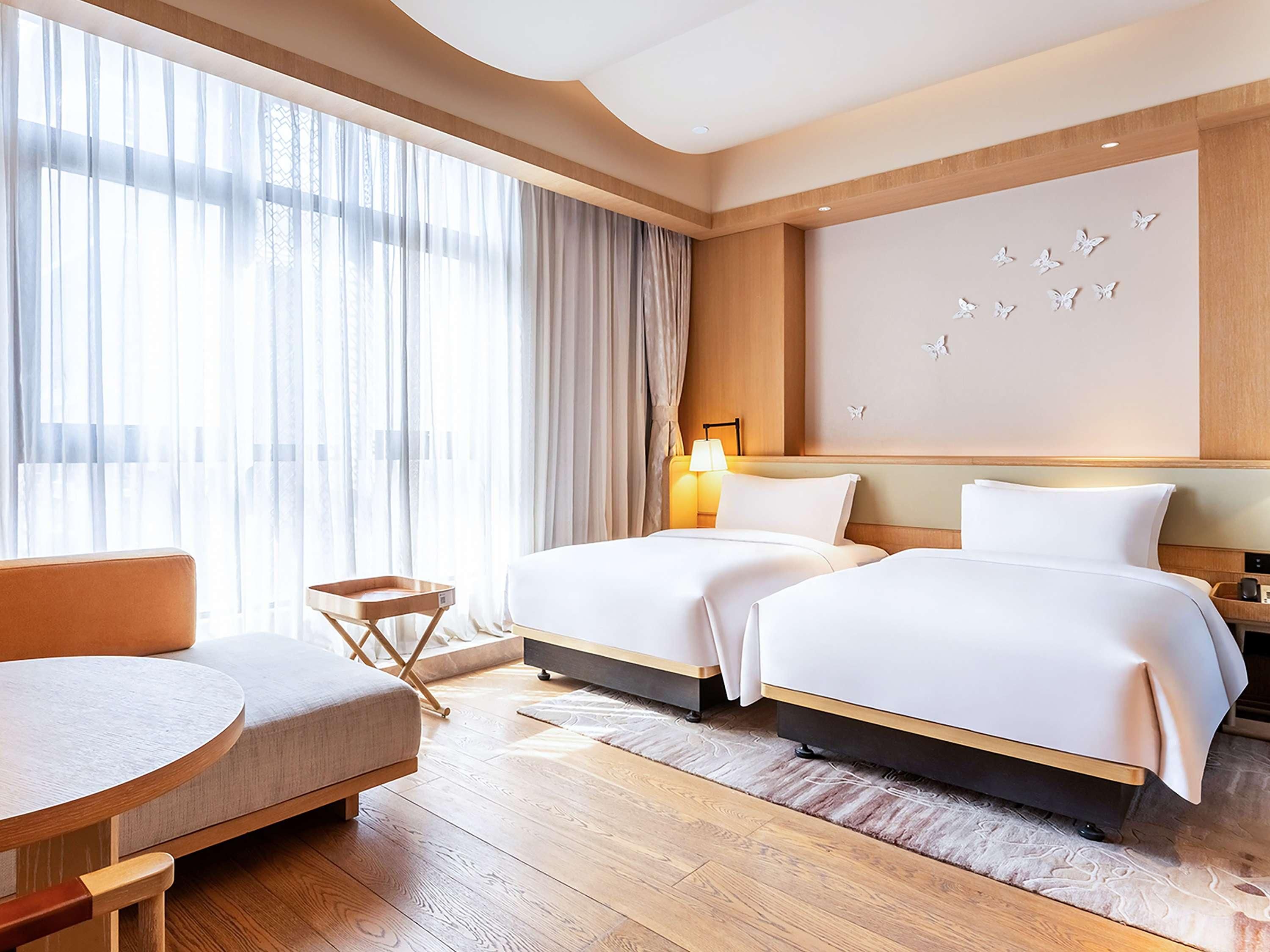 A hotel room with two beds, a sofa, a small table, and a large window with sheer curtains.