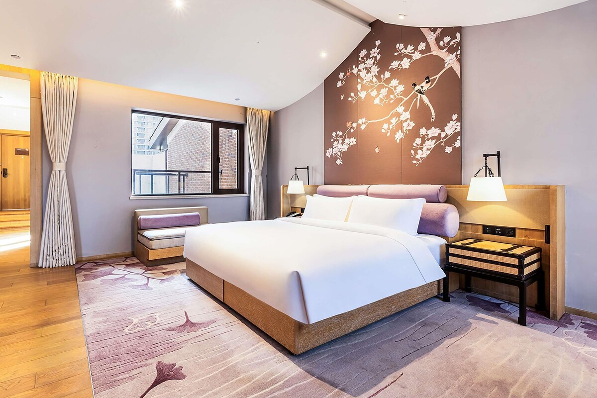 A modern hotel room with a large bed, a view of the city through the window, a decorative wall mural, and a bedside table with a lamp.