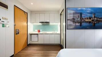 A hotel room with a wooden floor, a bed, a small kitchenette with a sink, and a cityscape view on the wall.