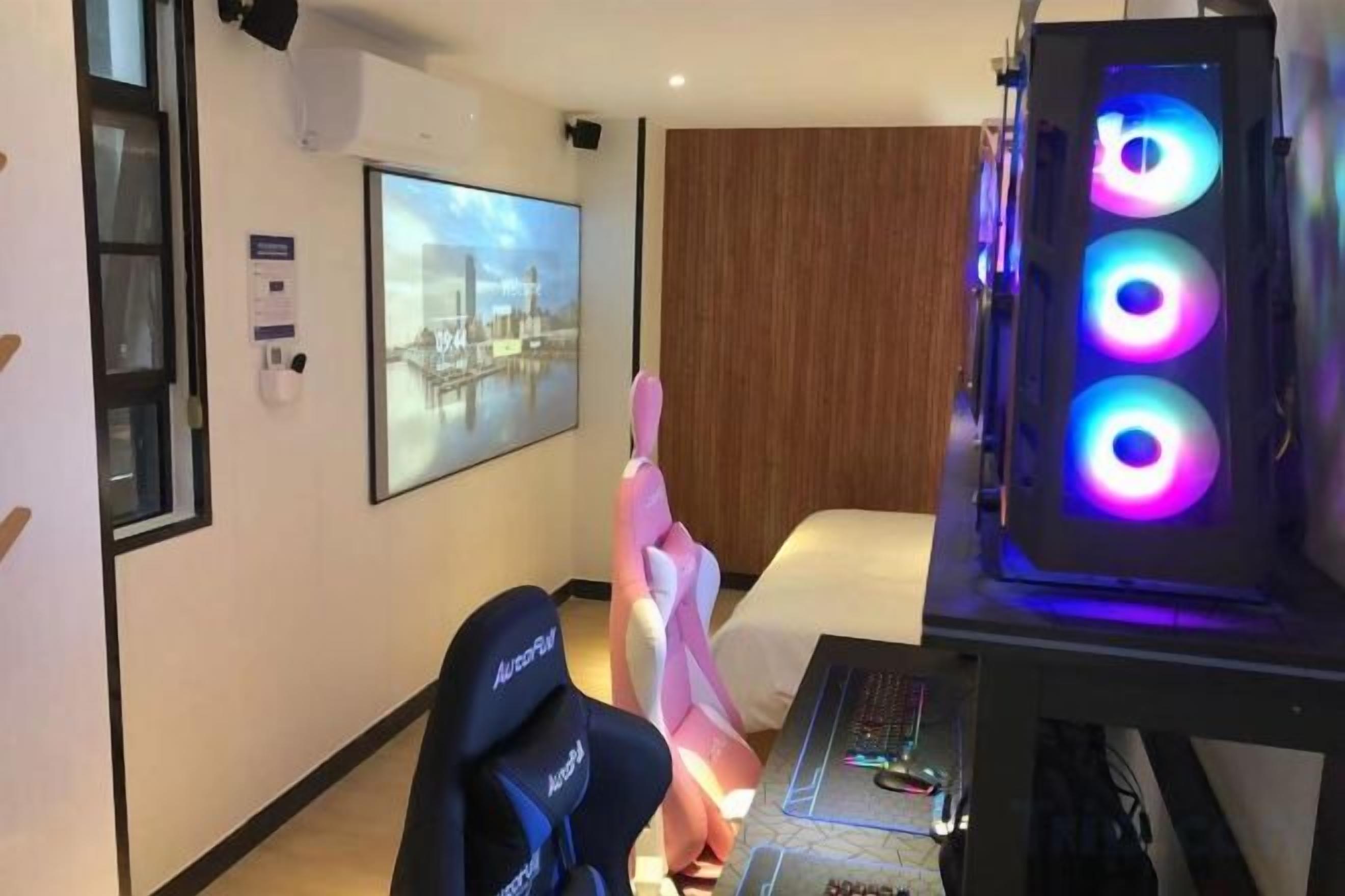 Une chambre d’hôtel équipée d’un lit, d’un bureau avec un ordinateur, d’un fauteuil de jeu et d’un haut-parleur fixé au mur.