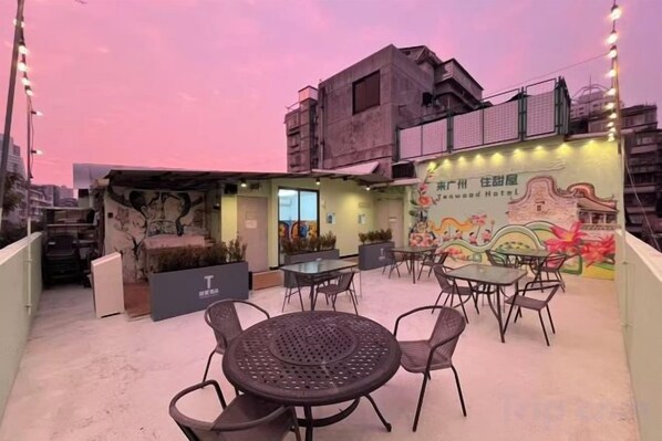 Un área de estar al aire libre con mesas y sillas, un mural y la fachada de un edificio con señalización.