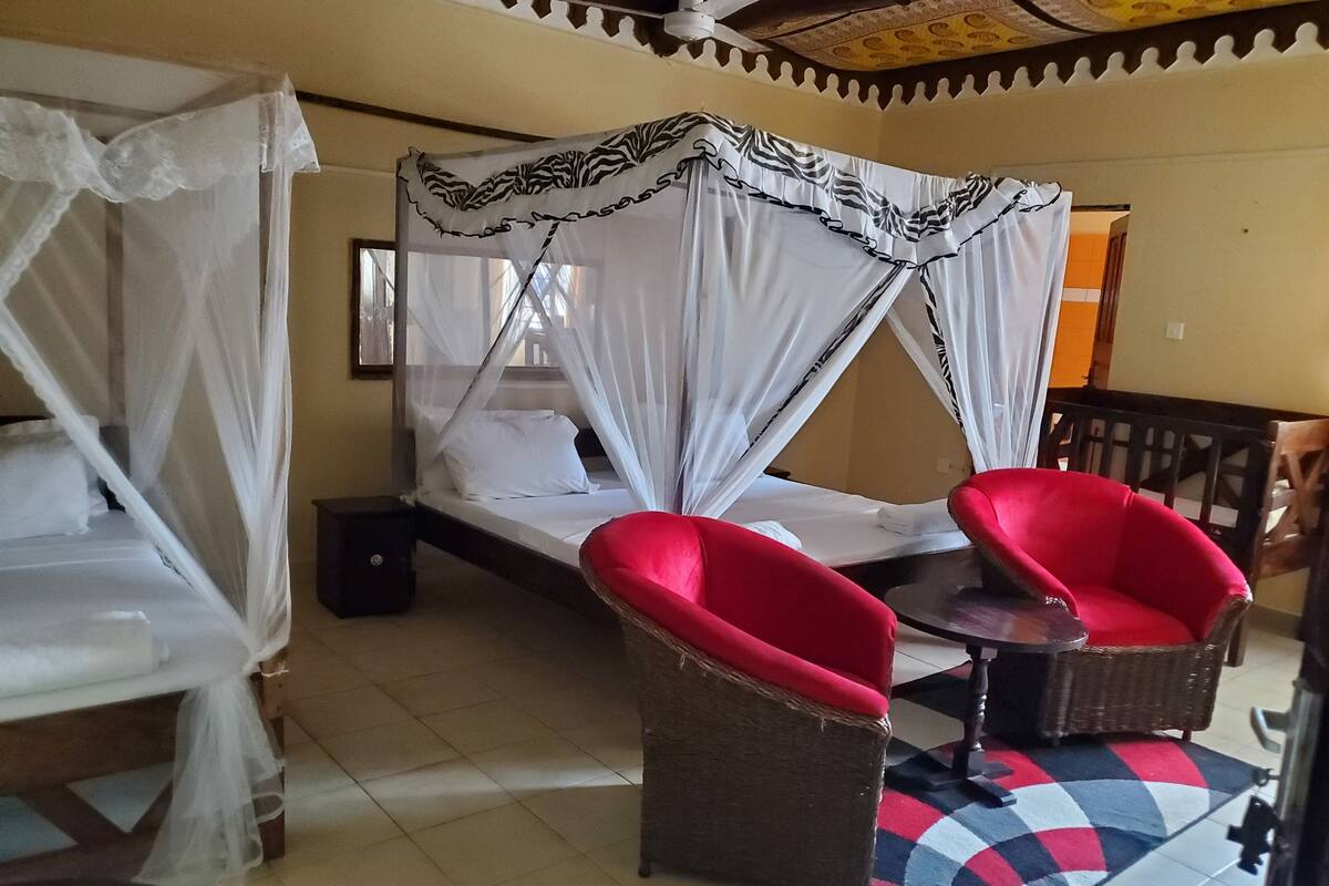 A room with a bed under a canopy, two red chairs, a small table, and a patterned rug.