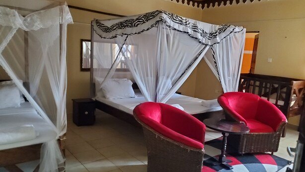 Una habitación con una cama bajo un dosel, dos sillas rojas, una mesita y una alfombra estampada.