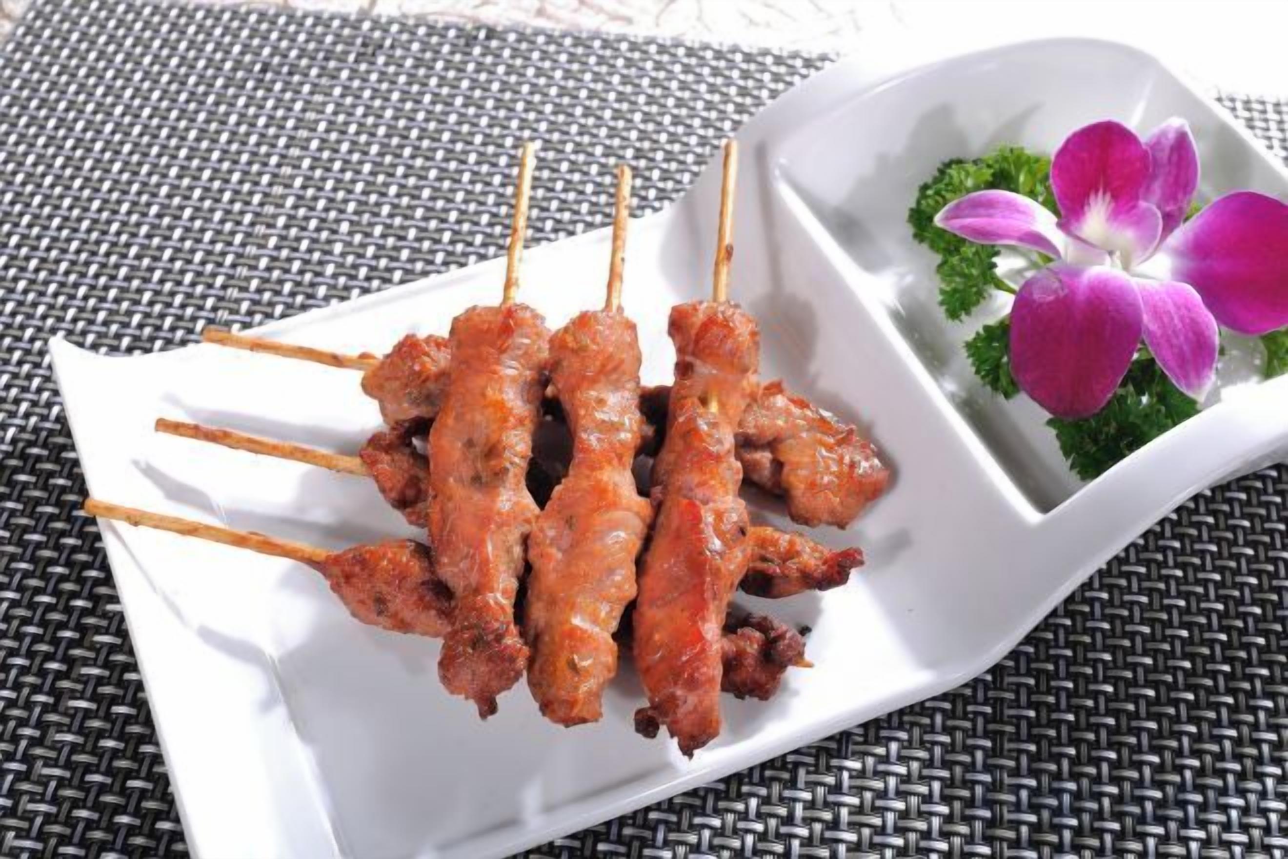 Une assiette rectangulaire blanche sur laquelle sont disposées des brochettes de viande, accompagnée d’un petit récipient contenant une fleur.