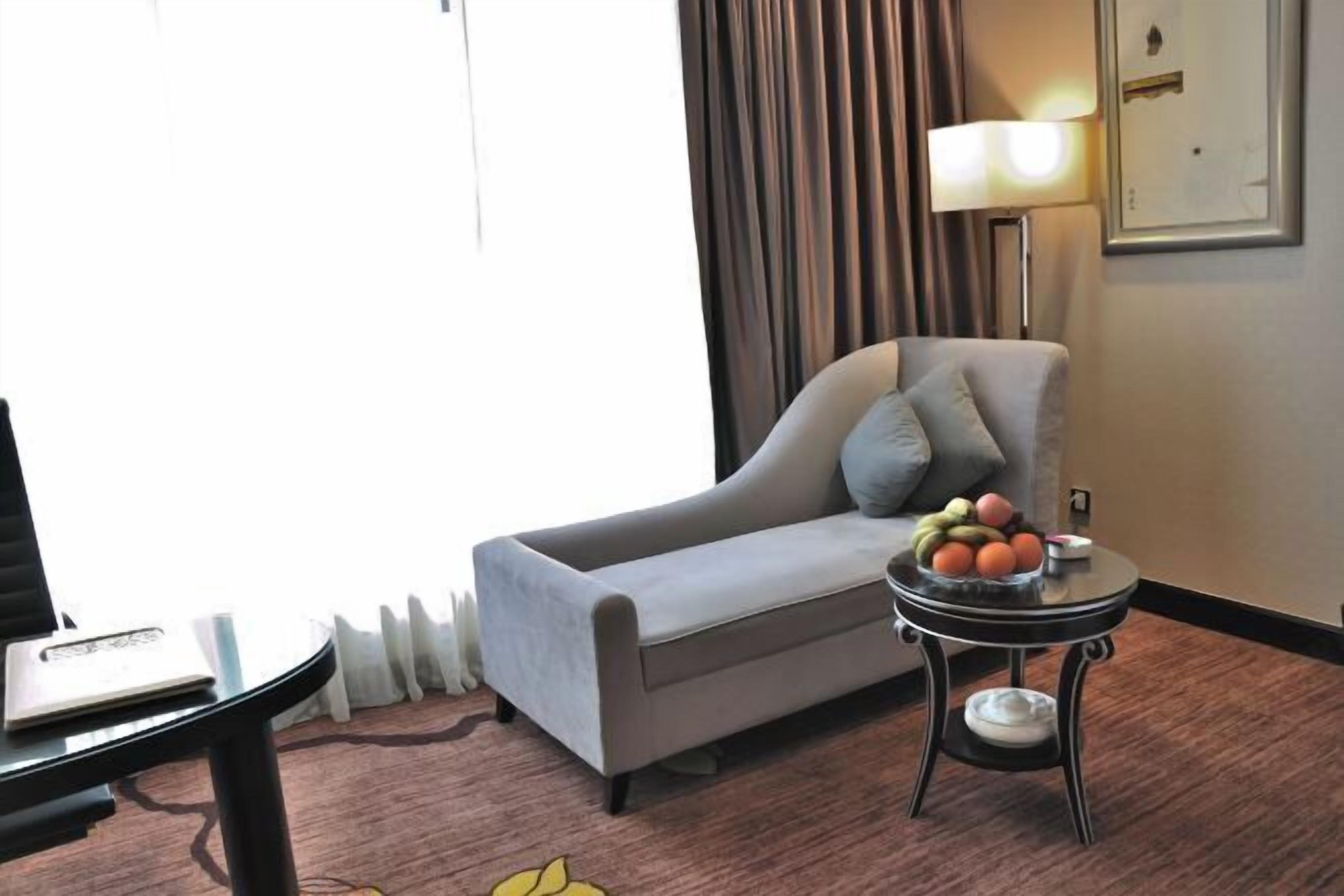 A hotel room with a grey sofa, a small round table with fruit, a lamp, and a laptop on a desk.