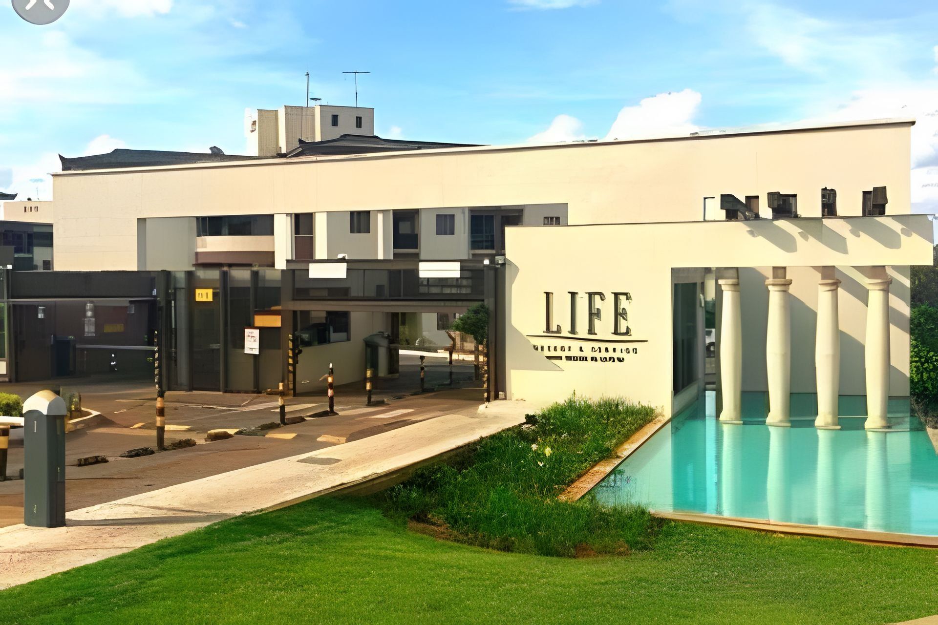 Um edifício moderno com piscina, entrada fechada por portão e um letreiro que diz 'LIFE'.