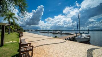 Um calçadão à beira-mar pavimentado, com bancos, fileira de palmeiras e um porto com barcos.