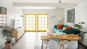 A bright living room with a wooden table, white chairs, a teal sofa with orange cushions, and a large window with yellow frames. - The Manzanita Cottage (Sebastopol)