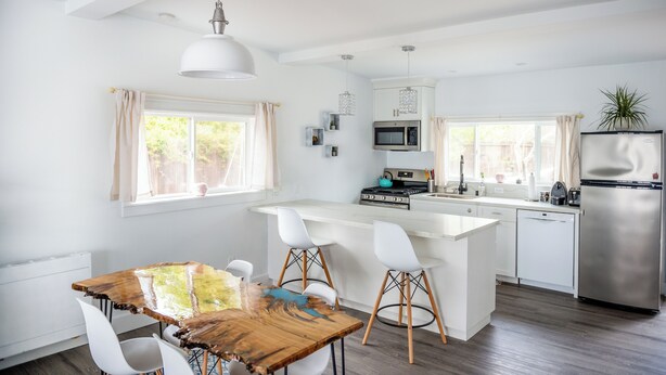 A modern kitchen with white cabinetry, a central island with bar stools, a stainless steel refrigerator, and a hanging pendant light. - The Manzanita Cottage (Sebastopol)