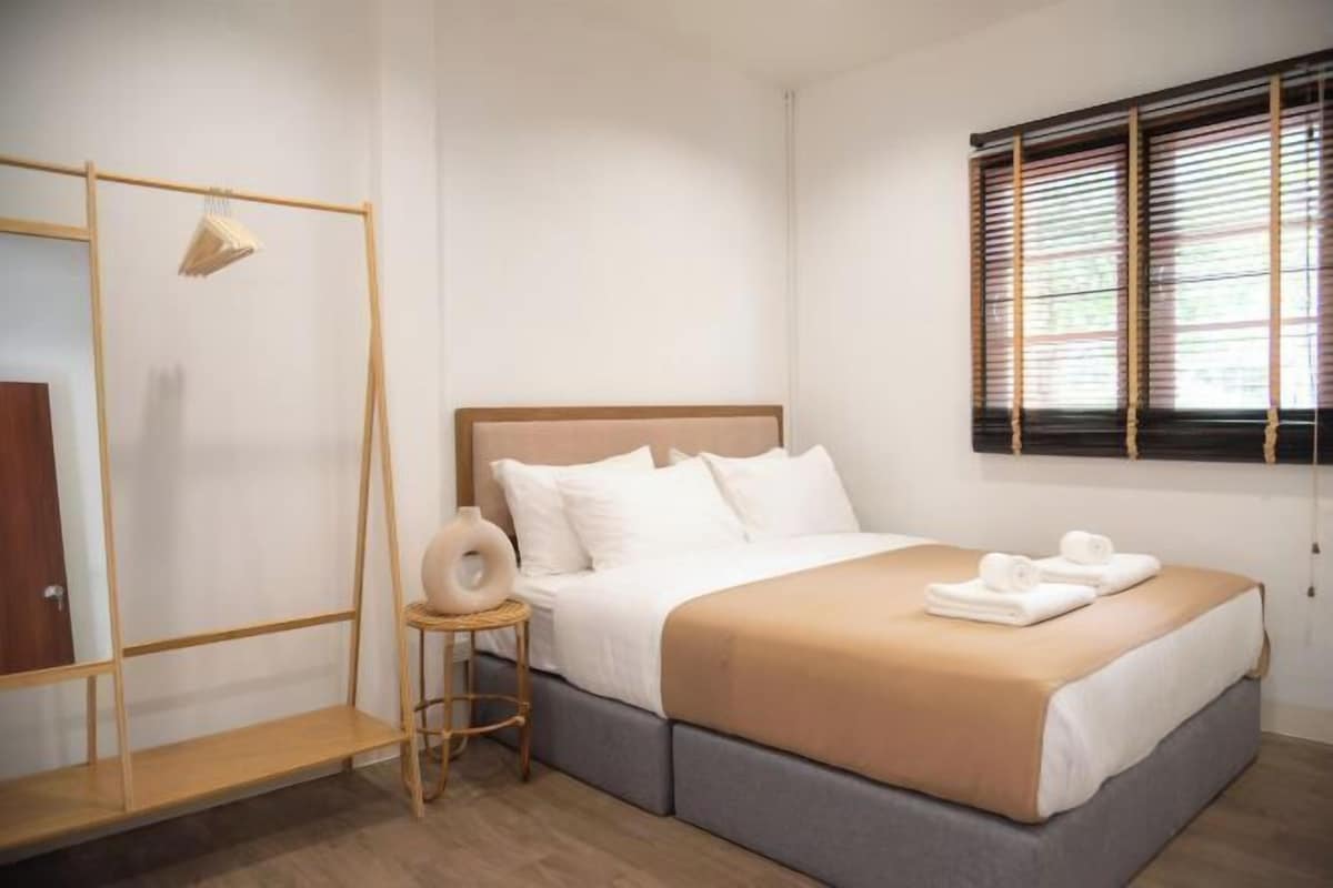 A neatly arranged bedroom with a bed, a wooden shelf, a small stool, and a window with blinds.