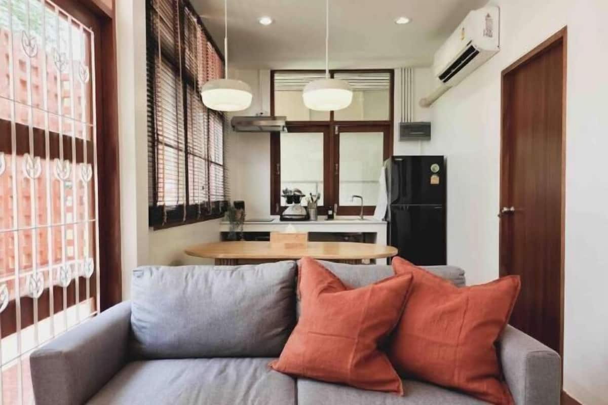 A living room with a sofa and two pillows, a dining area with a table and chairs, and a kitchen with appliances and a refrigerator.