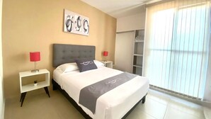 Une chambre d’hôtel comprenant un lit, deux tables de chevet, une petite table, une armoire et une fenêtre avec des stores.