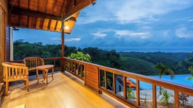 A wooden deck with two chairs and a table, overlooking a lush landscape and a pool. - Nusa Penida Pini Sentana (Bali)
