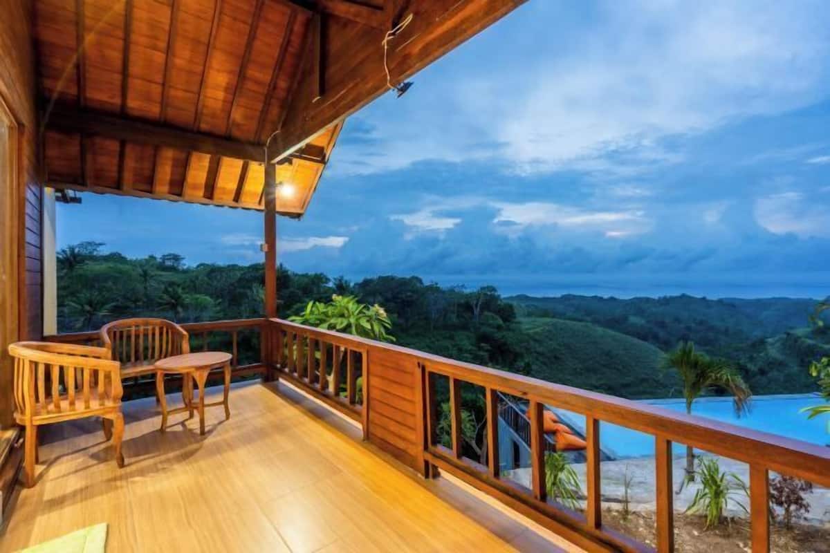 A wooden deck with two chairs and a table, overlooking a lush landscape and a pool.
