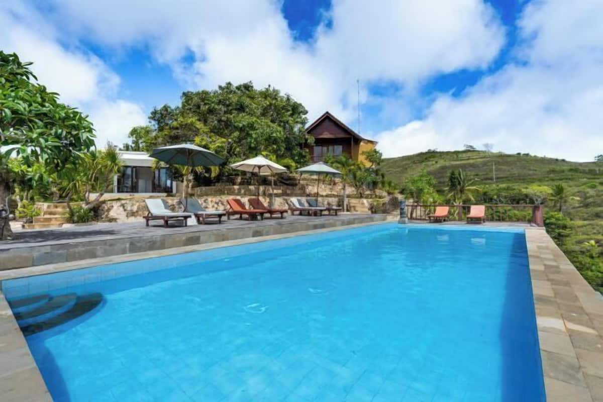 A swimming pool with lounge chairs and umbrellas, surrounded by greenery and a hill in the background.