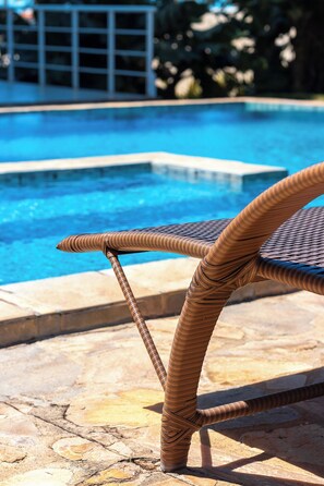 A wicker lounge chair by a swimming pool with a stone patio.