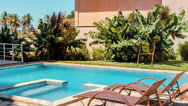 A swimming pool with clear blue water, surrounded by tropical plants and a building in the background. - 2 suítes térreo - próximo da piscina (Aquiraz)