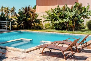 A swimming pool with clear blue water, surrounded by tropical plants and a building in the background. - 2 suítes térreo - próximo da piscina (Aquiraz)