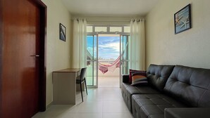 A living room with a sofa, a small table, and a chair. There is a door on the left and a window with curtains on the right. - 2 suítes térreo - próximo da piscina (Aquiraz)