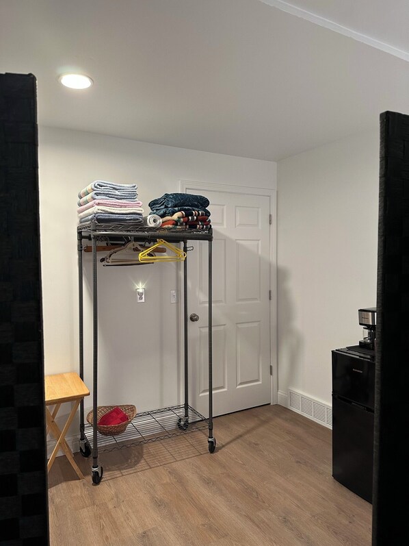 A room with a metal rack holding folded towels and blankets, a small wooden stool, a basket with a red item, a coffee machine, and a door.