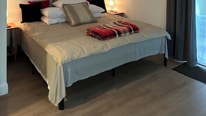 Una cama bien hecha con una colcha a rayas y una manta roja y blanca, una lámpara de noche y una ventana con cortinas.
