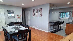 A modern dining area with a wooden table and chairs, a kitchen with bar stools, and a large window with curtains. - Spacious 4-bedroom house in charming Des Plaines with AC, WiFi (Des Plaines)