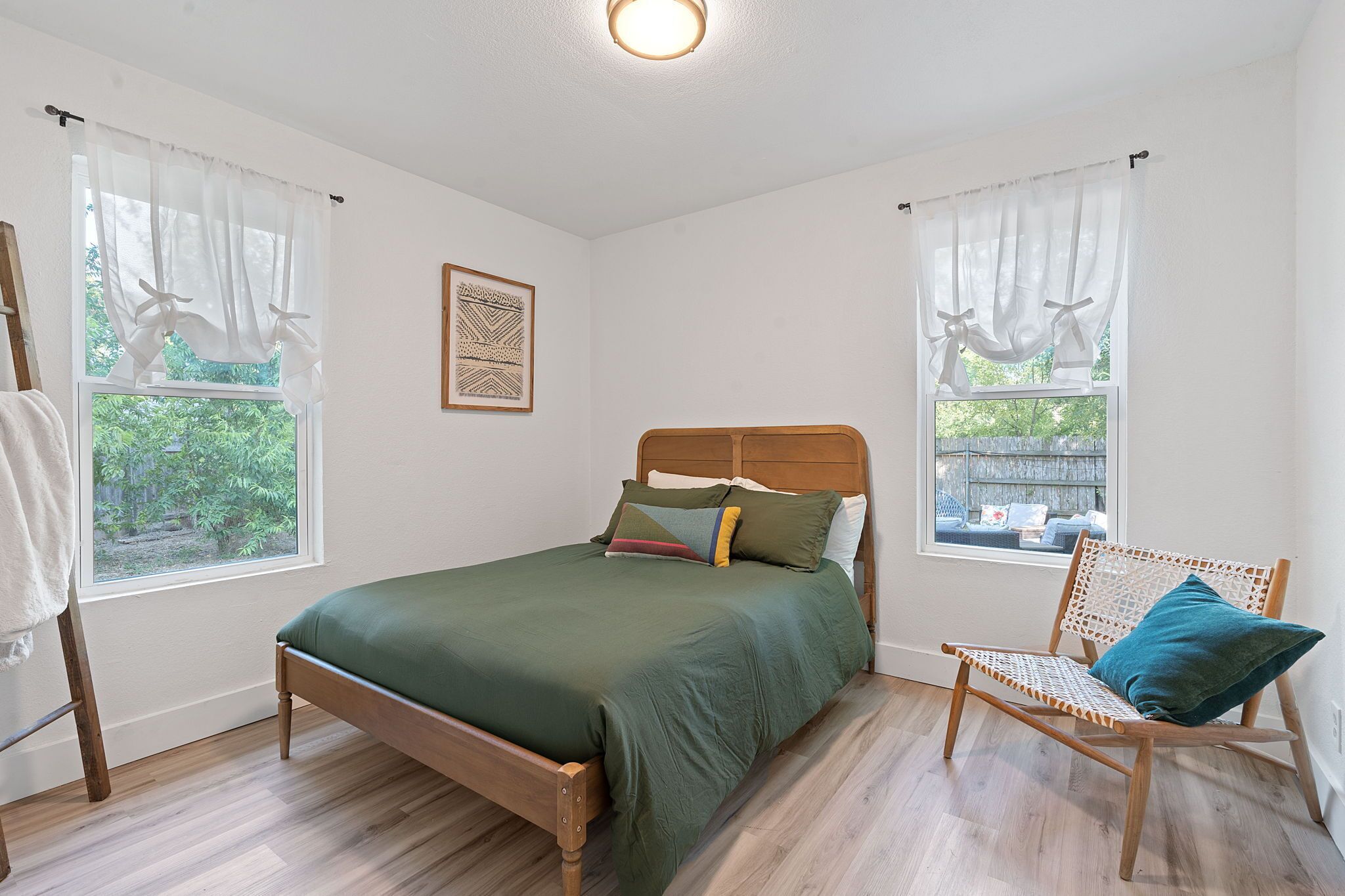 A bedroom with a wooden bed, two windows with sheer curtains, a wooden chair with a cushion, and a framed artwork on the wall.