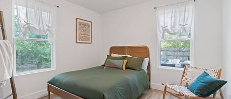 A bedroom with a wooden bed, two windows with sheer curtains, a wooden chair with a cushion, and a framed artwork on the wall.