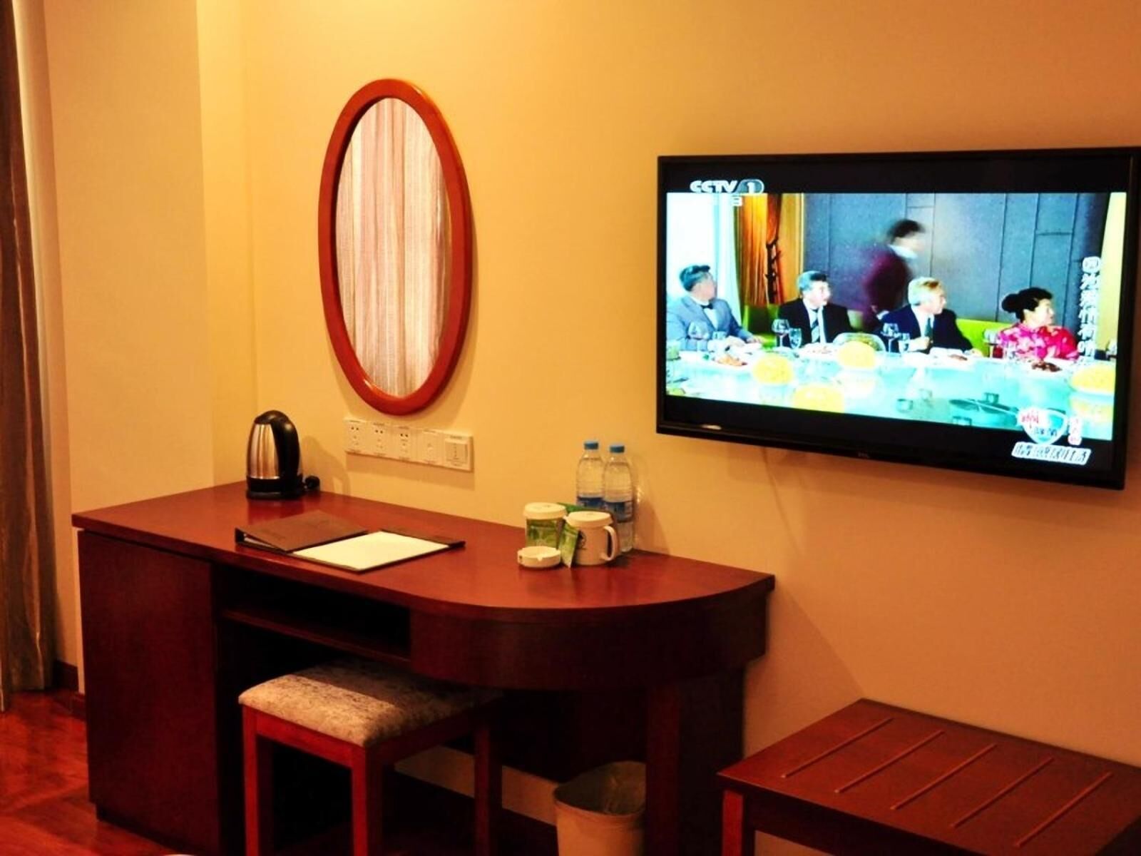 A hotel room with a wooden desk, a television mounted on the wall, a mirror, and a small seating area.