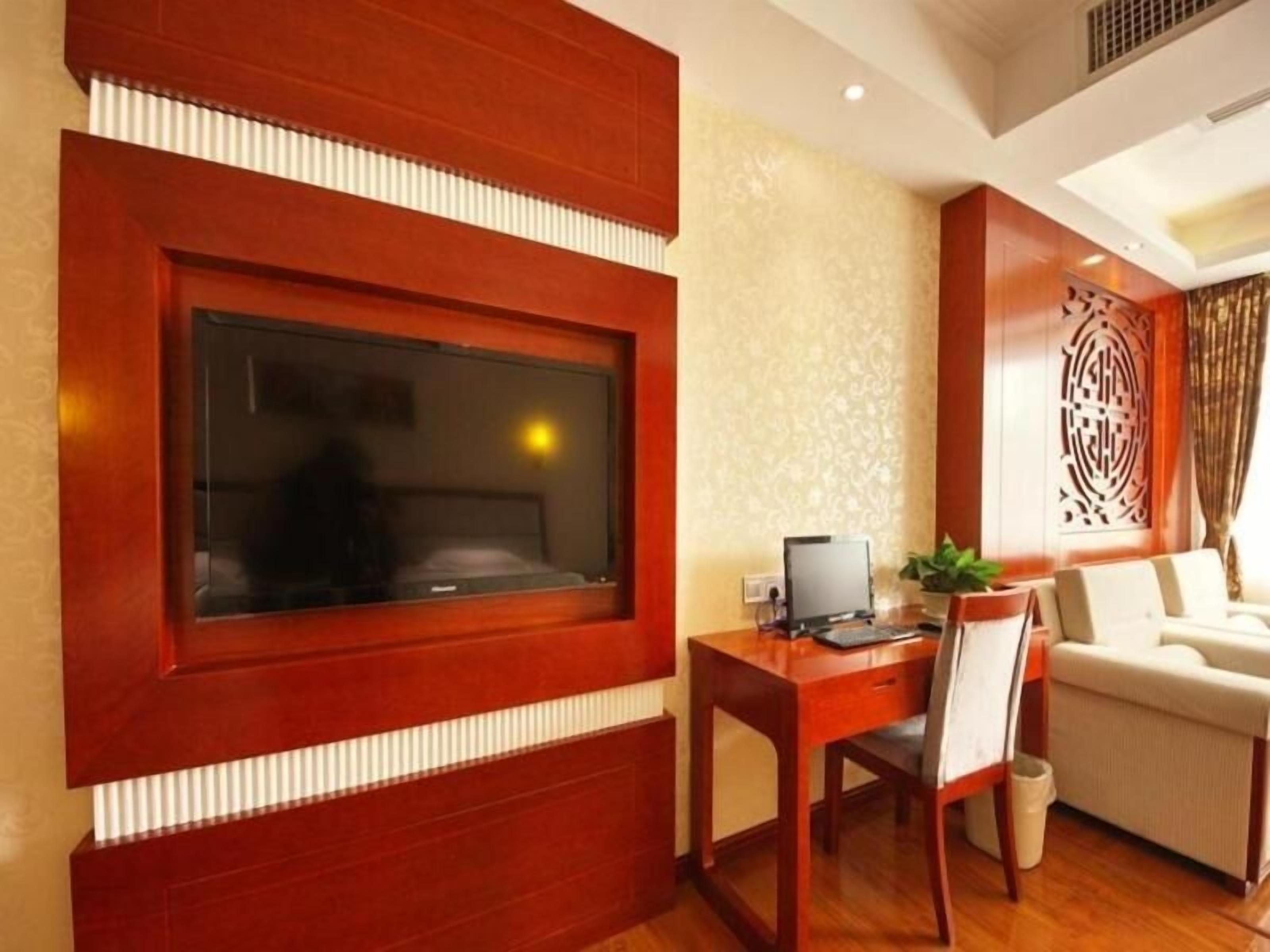 A hotel room with a large TV mounted on a wooden panel, a desk with a computer, a chair, and a sofa.