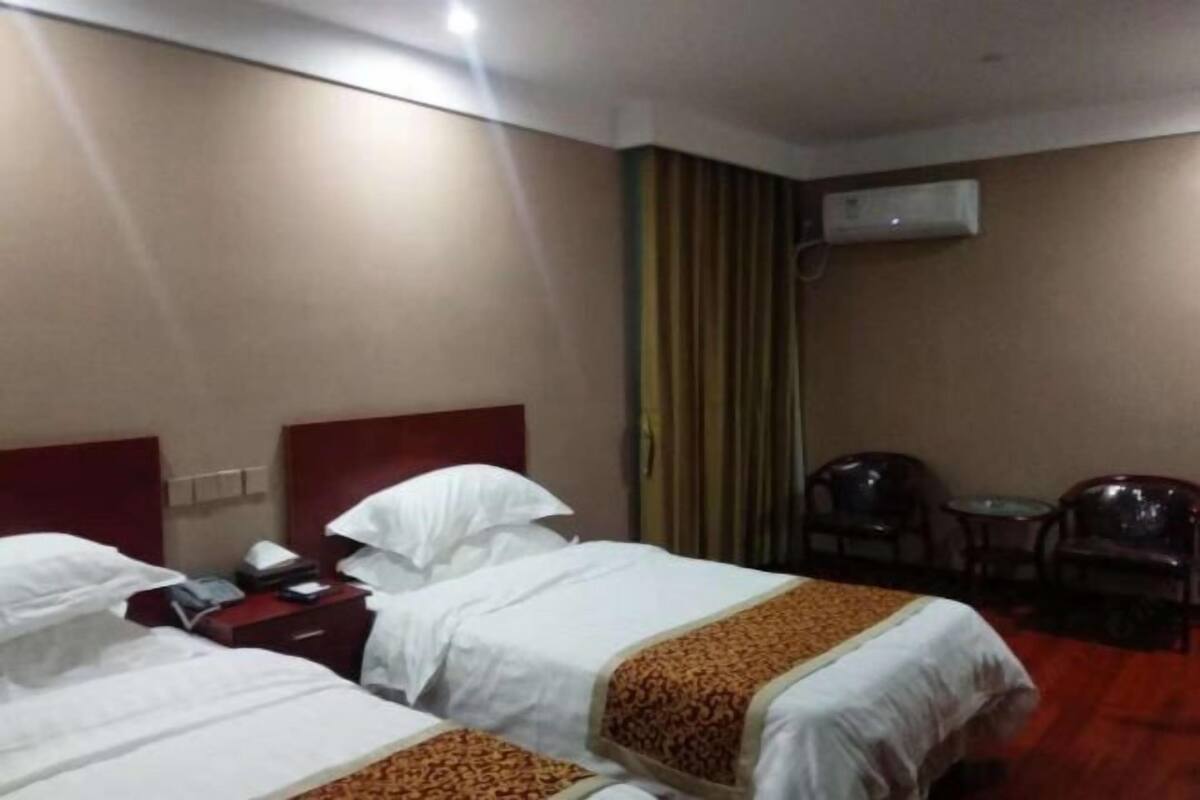 Habitación de hotel con dos camas, una mesita de noche con lámpara, un equipo de aire acondicionado en la pared y una pequeña mesa con sillas.