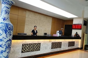 Un mostrador de recepción con una recepcionista, un jarrón decorativo grande y una pantalla digital.