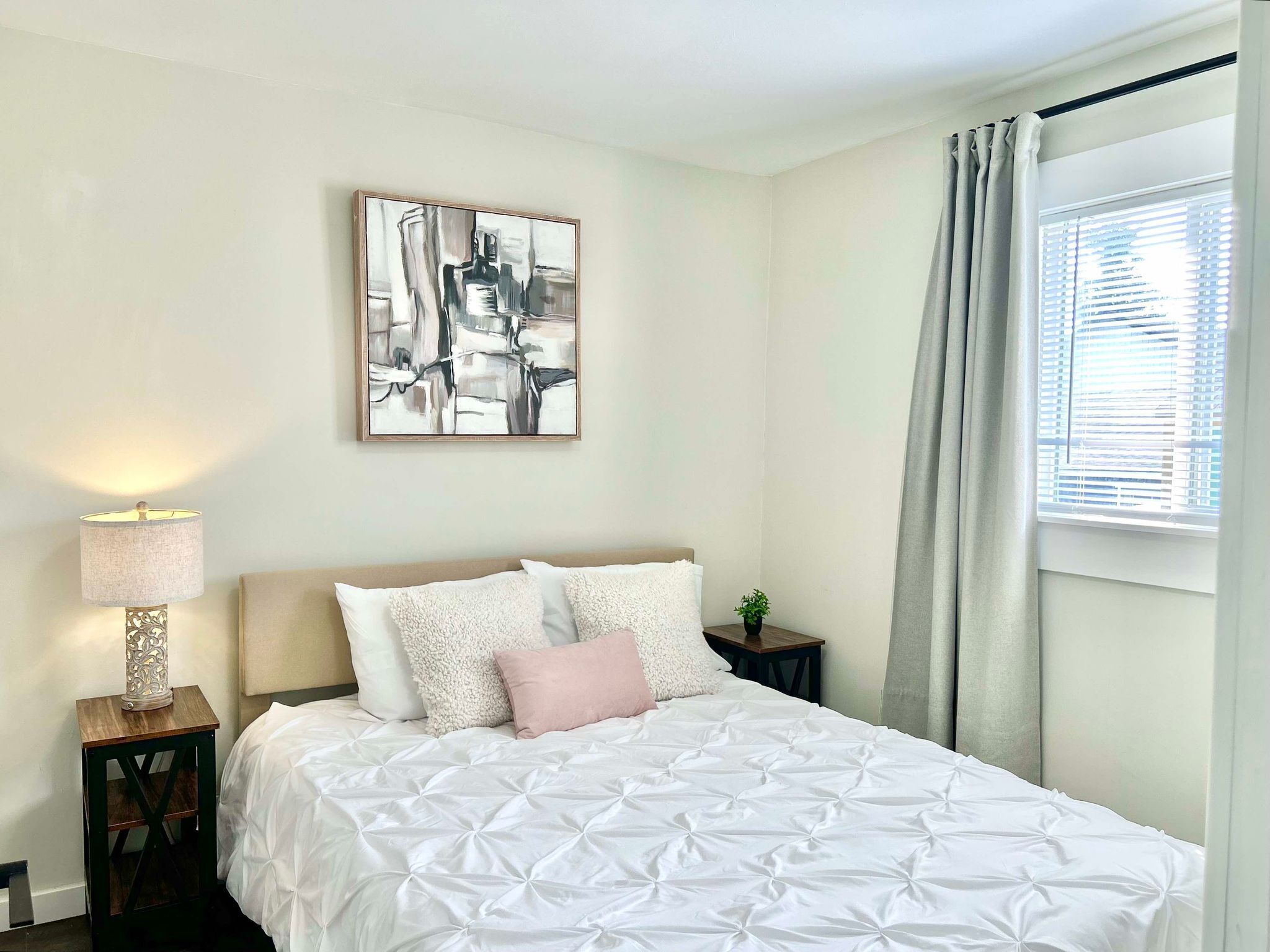 Une chambre à coucher bien rangée, avec un lit, une table de chevet sur laquelle se trouve une lampe, une petite plante en pot et une fenêtre avec des rideaux.