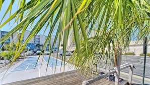 A wooden deck with a railing, palm trees, and a parking lot with cars and buildings in the background.