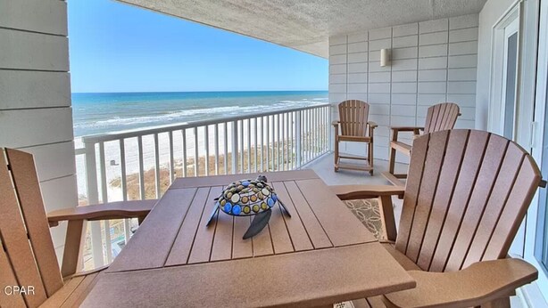 A balcony with a view of the beach, wooden chairs, and a table with a decorative bowl.
