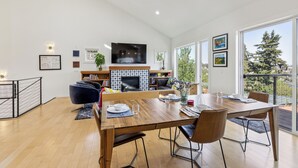 A modern dining area with a wooden table set for four, a fireplace, and a large window offering a view of trees. - 4-Bd w/ AC, Walk to Light Rail, Yard, VIEW Dog OK! (Seattle)