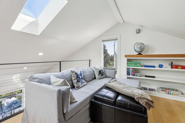 A modern loft bedroom with a skylight, a grey sofa with decorative pillows, a black leather ottoman, and a bookshelf with various items. - 4-Bd w/ AC, Walk to Light Rail, Yard, VIEW Dog OK! (Seattle)