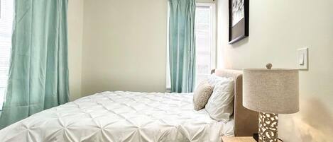 A neatly made bed with a white duvet, a bedside lamp with a patterned shade, and a framed picture on the wall.