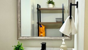 A bathroom with a white sink, a black faucet, a mirror with a wooden frame, a shelf with a potted plant, a yellow bottle, and a white bottle. - Stylish Deluxe Kitchen /King Bed/Laundry/Free Parking (Renton)