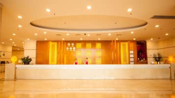 A hotel reception area with a reception desk, two staff members, and a decorative wall with framed items.
