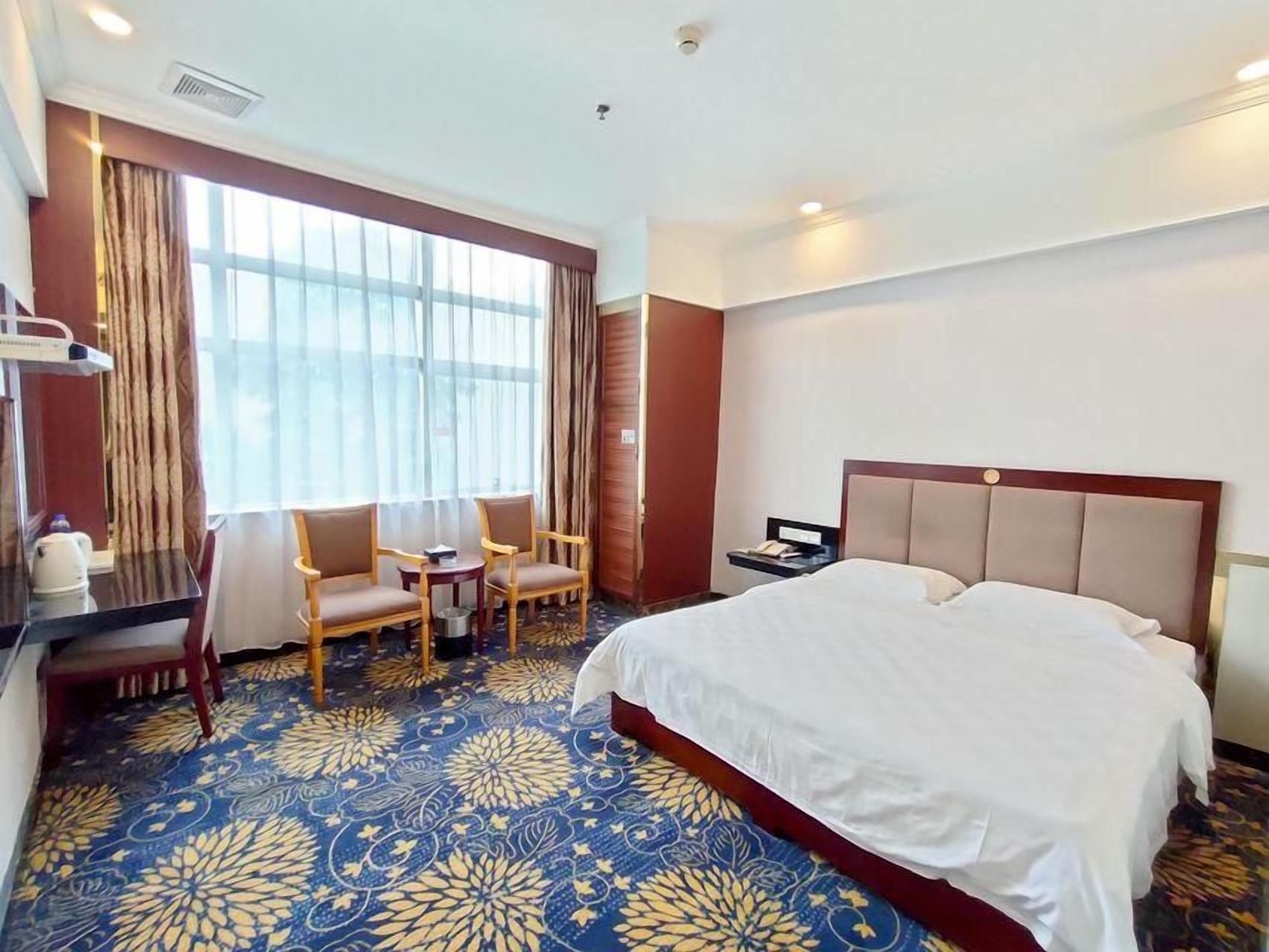 Una habitación de hotel con una cama grande, un escritorio con silla, una mesita y una ventana con cortinas.