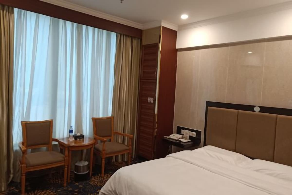 Une chambre d’hôtel avec un lit, deux chaises, une petite table, un téléphone et une fenêtre avec des rideaux.