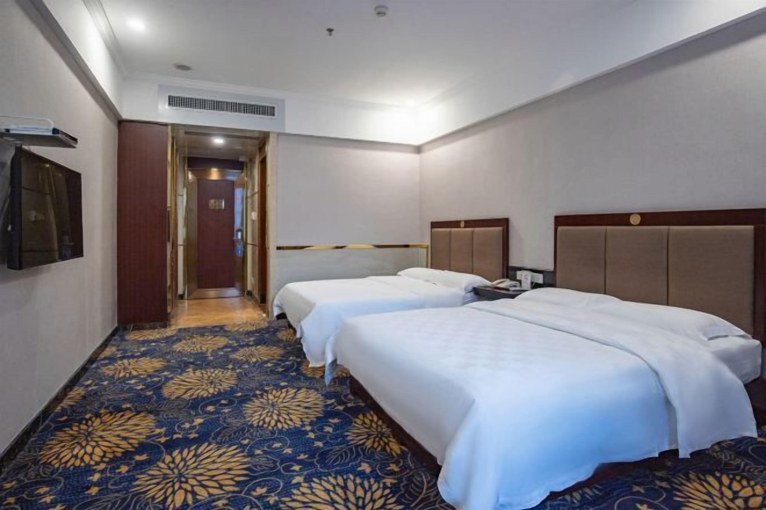 A hotel room with two beds, a patterned carpet, a wooden headboard, and a door leading to another room.