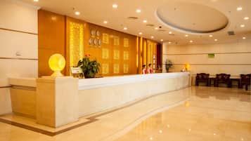 Uma área de recepção de hotel com piso de mármore, balcão de recepção e uma parede com relógios e painéis decorativos.