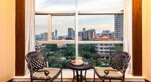 A room with two wicker chairs and a small round table, a large window with a cityscape view, and a curtain on the left.