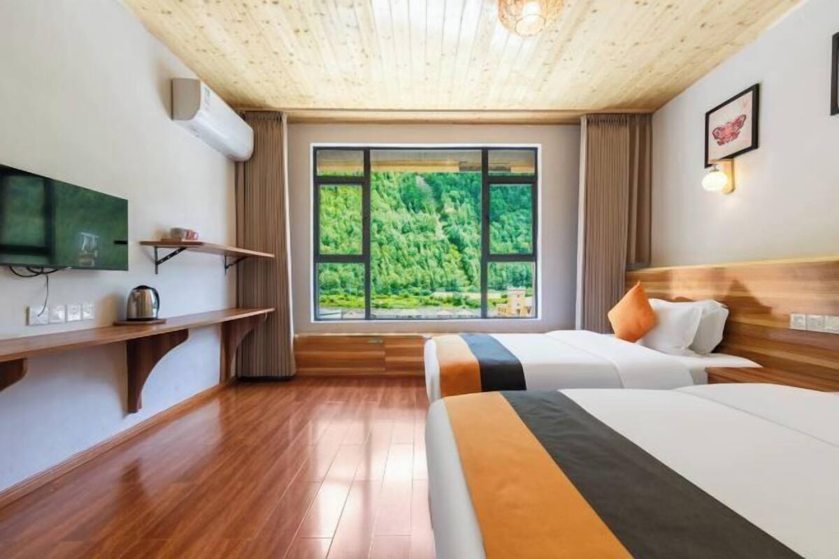 A hotel room with a wooden ceiling, a large window with a forest view, a bed, a bedside table, and a mounted TV.